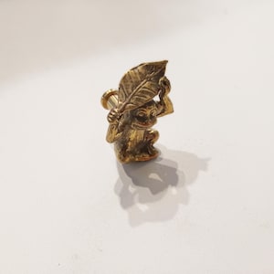 May include: A gold monkey figurine holding a leaf over its head. The monkey is sitting on a small base.