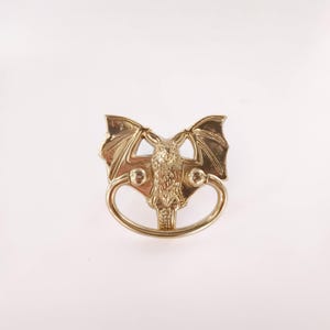 以下が含まれることがあります： 金色のコウモリ型のドアノッカー。ノッカーは、広げられた翼を持つ詳細なコウモリと、その下の円形のハンドルが特徴です。デザインは対称的で、装飾的です。