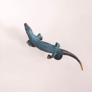 Può includere: Scultura di lucertola in metallo blu-verde con corpo strutturato e lunga coda. La lucertola ha il ventre e le zampe color bronzo. Un oggetto decorativo, forse per l'arredamento della casa.