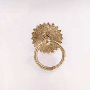 Op de afbeelding: Een goudkleurige bloemvormige trekker met een ringhandvat. De trekker heeft gedetailleerde bloemblad- en middendesigns, wat een decoratief tintje geeft. De ringhandgreep is bevestigd aan het midden van de zonnebloem.