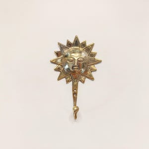 May include: A gold metal sun-shaped hook with a face in the center. The hook is attached to a wall.