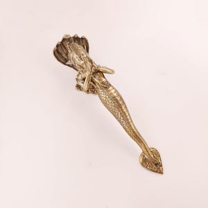 May include: A brass-colored decorative door handle featuring a detailed sculpture of a figure with a serpent hood. The figure has arms crossed and a scaled body. The handle has a heart-shaped end.