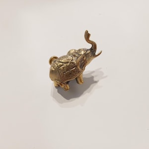 May include: A small, gold-coloured metal elephant figurine with intricate details. The elephant's trunk is raised and its head is tilted back.