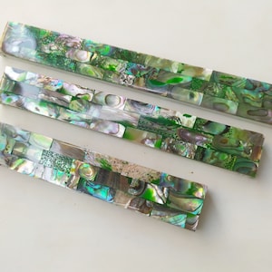 May include: Three green, iridescent, rectangular pieces of abalone shell, arranged in descending size order. The shell is made up of small, rectangular pieces of abalone, creating a mosaic-like pattern.