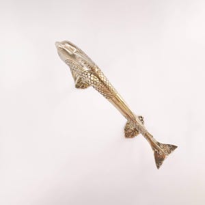 May include: A decorative, silver-toned door handle in the shape of a fish. The handle features detailed scales, fins, and a tail. The fish-shaped handle is mounted on a white background.
