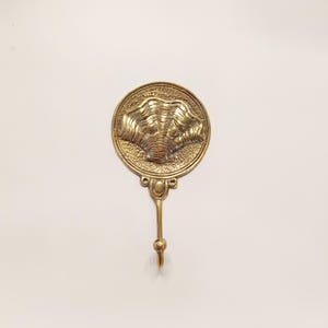 May include: A gold-toned metal hook with a decorative round top featuring a raised design of two stylized shells. The hook is attached to a wall.