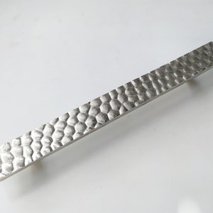 May include: A silver metal cabinet pull with a hammered texture. The pull is rectangular in shape and has a smooth, rounded edge.