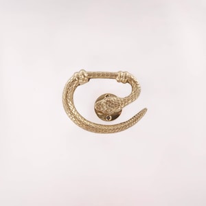 May include: A brass-colored door handle in the shape of a snake. The handle features a detailed snake head and body, with a textured scale pattern. The handle is designed to be mounted on a door and adds a unique decorative element.