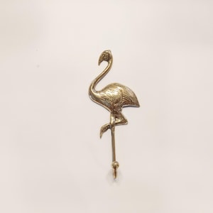 May include: A gold metal flamingo hook. The flamingo is standing on one leg with its head bent down. The hook is attached to the flamingo's leg.