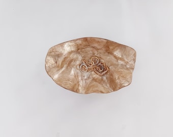 Puede incluir: Un plato de joyería decorativo, de forma libre, iridiscente y de color bronceado. Varias piezas de joyería de oro y plata descansan dentro del plato. El plato tiene una apariencia texturizada y orgánica y se asienta sobre un fondo blanco.