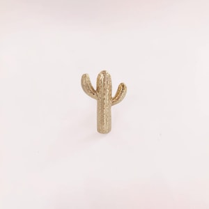 Può includere: Un oggetto decorativo a forma di cactus color oro. Il cactus ha un fusto verticale con tre braccia curve. L'oggetto è realizzato in materiale metallico ed è posto su uno sfondo bianco.