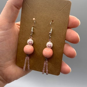 Puede incluir: Un par de pendientes colgantes con una cuenta blanca con una cara sonriente encima de una cuenta grande y redonda de color coral. Los pendientes tienen una borla de cuentas y ganchos plateados. Los pendientes se muestran en una tarjeta marrón.
