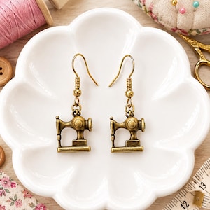 May include: A pair of antique brass-toned metal earrings shaped like vintage sewing machines. Each earring features a detailed miniature sewing machine charm suspended from a simple hook wire, showcasing a metallic finish with dark accents.