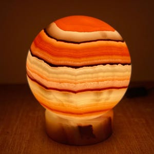 May include: A round, striped, orange and white onyx lamp with a base. The lamp is lit and glowing.