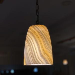 Può includere: Una lampada a sospensione con paralume in pietra naturale. Il paralume ha un motivo a strati giallo caldo e bianco. La luce è appesa a una catena nera.