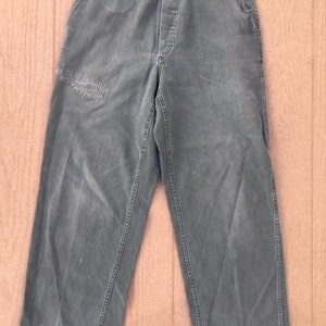 Può includere: Un paio di pantaloni da lavoro verdi con chiusura a bottone e diverse tasche. I pantaloni hanno un aspetto usurato con alcuni buchi e strappi.