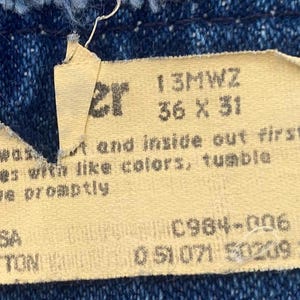以下が含まれることがあります： Wranglerジーンズのタグのクローズアップ。タグはベージュで、黒い文字で「Wrangler」、サイズ36 x 31、洗濯表示などが記載されています。タグは、100％コットンの青いデニム生地に縫い付けられています。