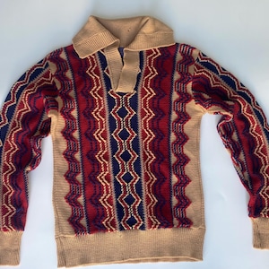 May include: A long-sleeve knit sweater featuring a tan collar and cuffs with a repeating vertical pattern of red, navy blue, and tan zigzags and diamond shapes. The pullover design includes a notched neckline and a textured knit construction.