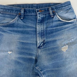 以下が含まれることがあります： ダメージ加工が施された、明るいブルーデニムのジーンズのクローズアップ。ジーンズは、クラシックな5ポケットのデザイン、ボタンとジッパーの開閉部、いくつかの小さな破れや色あせた部分を含む、目に見える摩耗と損傷があります。