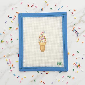 May include: A needlepoint design featuring a pixelated ice cream cone with colorful sprinkles. The design is set against a white background, framed by a blue border with green stitching. The initials "AC" are in the lower right corner. Sprinkles are scattered around the frame.