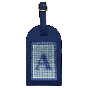 Puede incluir: Una etiqueta de equipaje azul marino con hebilla y correa doradas. La etiqueta presenta la letra "A" bordada en azul claro y blanco sobre un parche rectangular. La etiqueta est&aacute; hecha de un material texturizado.