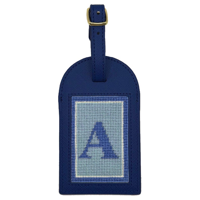 Puede incluir: Una etiqueta de equipaje azul marino con hebilla y correa doradas. La etiqueta presenta la letra "A" bordada en azul claro y blanco sobre un parche rectangular. La etiqueta est&aacute; hecha de un material texturizado.