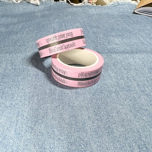 May include: Two pink rolls of tape with black stripes and text in a foreign language. The tape is on a blue denim background.