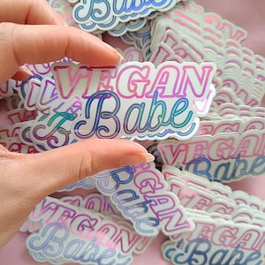 以下が含まれることがあります： ピンク、ブルー、パープルのグラデーションで「Vegan Babe」というテキストが書かれた、虹色のホログラフィックステッカーのスタック。ステッカーはピンクの背景の上にあります。
