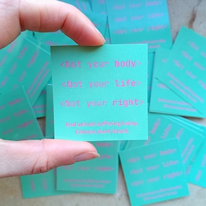 Peut inclure: Une main tient une carte carrée turquoise avec le texte "<Not your body>", "<Not your life>" et "<Not your right>" en rose. La carte indique également "End animal suffering today. Choose plant based." D'autres cartes sont visibles en arrière-plan.