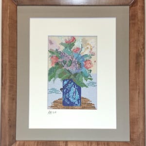May include: Framed needlepoint art featuring a floral arrangement in a blue vase. The flowers are a mix of pink, red, and blue, with green leaves. The vase has a blue pattern. The artwork is framed in a wooden frame.