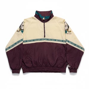 Könnte beinhalten: Vintage Anaheim Mighty Ducks Starter Jacke. Die Jacke hat einen cremefarbenen oberen Teil und einen weinroten unteren Teil mit einem türkis-weißen Kragen. Das Mighty Ducks-Logo befindet sich auf den Ärmeln. Die Jacke hat einen halben Reißverschluss und einen gerippten Saum.