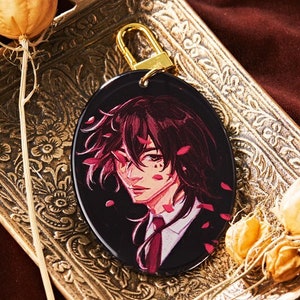 May include: A gold-toned oval keychain with a black and white illustration of a person with pink hair and a red tie. The keychain is on a gold tray with a floral design and a dried flower.