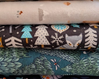 May include: A stack of five folded fabric bolts featuring various nature-themed prints. The patterns include a beige moose print, a light gray mountain scene, a dark forest with a fox and snowflakes, a teal pine tree forest, and a dark blue rabbit print.