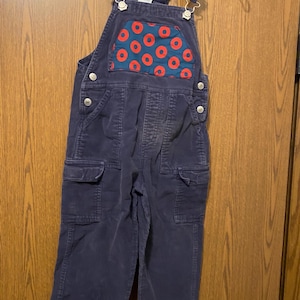 May include: Navy blue corduroy overalls for children. The bib pocket features a red and blue pattern. The overalls have adjustable straps, side pockets, and button closures. The garment is hanging on a clear plastic hanger.