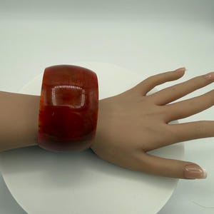 May include: A wide, reddish-brown bangle bracelet. The bracelet is worn on a wrist and has a smooth, polished surface. The color transitions from a lighter orange-red to a darker brown. The bracelet is a bold statement piece.
