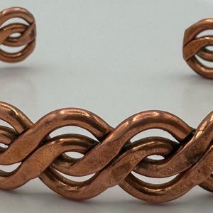 May include: A copper cuff bracelet with a woven design. The bracelet features a series of interconnected loops, creating a textured and open pattern. The metal has a warm, reddish-brown hue, and the bracelet is open at the ends.