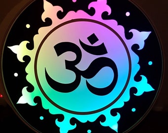 May include: A circular wall decor with a black background and a colorful om symbol in the center. The om symbol is surrounded by a decorative design with a rainbow gradient.