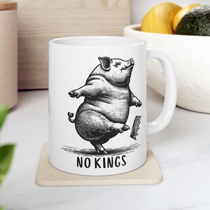May include: White ceramic mug featuring a detailed black and white illustration of a dancing pig. The mug has the text "NO KINGS" printed in a bold font at the bottom. The mug is on a beige coaster.