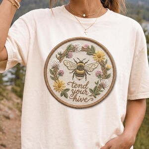 May include: A cream-colored short-sleeve t-shirt featuring a circular embroidered patch on the chest. The patch displays a central bee surrounded by a floral wreath and the text "tend your hive" in a brown script font.