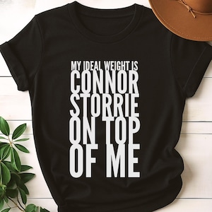 May include: Black t-shirt with white text that reads "MY IDEAL WEIGHT IS CONNOR STORRIE ON TOP OF ME." A brown hat is visible in the upper right corner. The word "BLACK" is printed in the lower right corner.