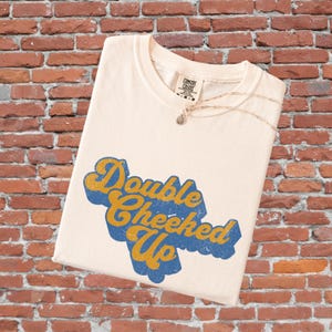 May include: Cream-colored t-shirt with the text "Double Cheeked Up" in blue and gold lettering. A gold necklace with a small pendant is draped across the shirt. The shirt is folded and displayed against a brick wall.