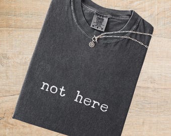 May include: A charcoal gray short-sleeve t-shirt with a crew neckline and the text "not here" printed in white on the chest. A silver-toned chain necklace with a circular pendant rests on the collar of the shirt, which features a Comfort Colors label.
