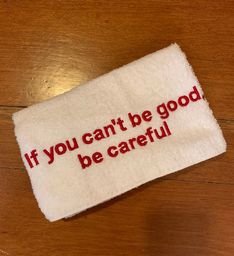 May include: White rectangular towel with the embroidered text "If you can't be good, be careful" in red. The towel is folded and lies on a wooden surface.