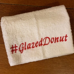 May include: White towel with red embroidered text that says "#GlazedDonut"
