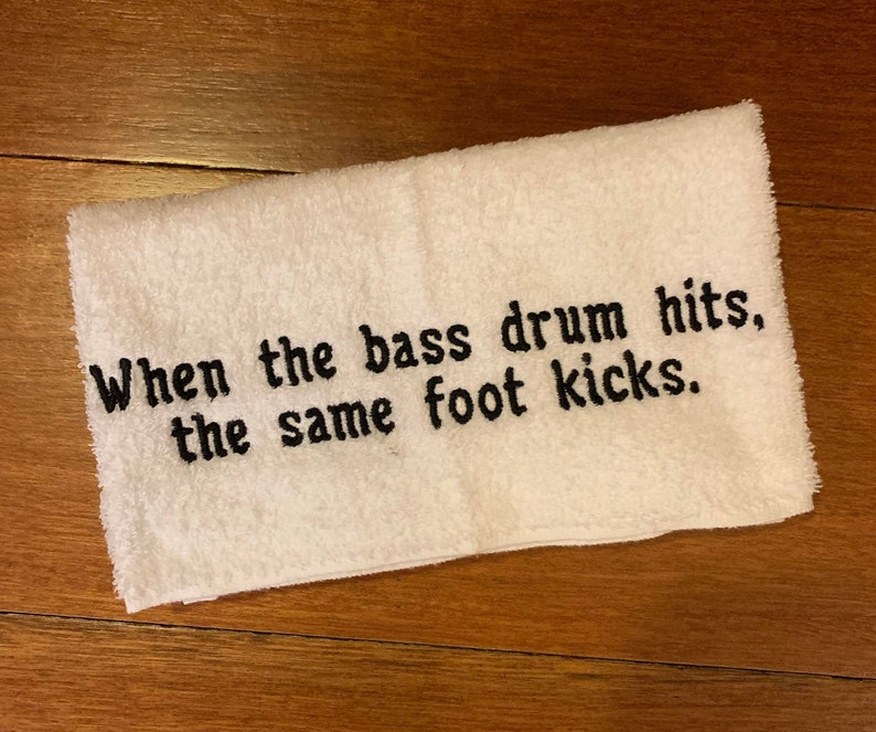 以下が含まれることがあります： 黒い刺繍のテキストが入った白い長方形のタオル。「When the bass drum hits, the same foot kicks.」タオルは柔らかく吸水性の高い素材でできており、バスルームやジムでの使用に最適です。テキストはタオルの中心に配置されています。