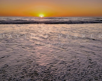 May include: A sunset over the ocean with a purple and orange sky. The waves are crashing on the shore, creating a foamy white edge. The water is calm and still, reflecting the colors of the sky. The image is taken from the beach, looking out at the ocean.
