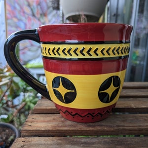May include: Red ceramic mug with a black handle and yellow and black geometric designs. The mug has two black circles with a white star in each circle.