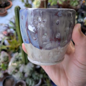 May include: A handmade ceramic cup with a floral pattern. The cup features a color gradient of blue, purple, and beige. The exterior has embossed flower and oval designs. The cup is held in a hand, with a blurred background of plants.