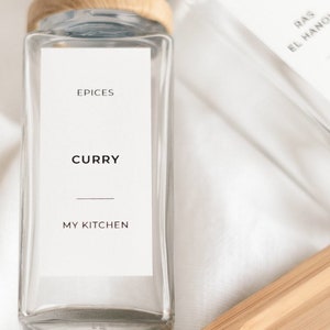 Può includere: Due barattoli di spezie in vetro con coperchi in legno ed etichette bianche. Le etichette riportano "Epices Curry MY KITCHEN" e "Epices RAS EL HANOUT MY KITCHEN". I barattoli sono su un vassoio di legno.