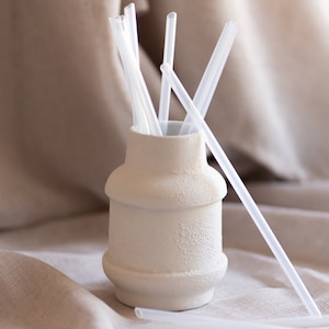 Könnte beinhalten: Eine weiße Keramikvase gefüllt mit durchsichtigen Plastikstrohhalmen. Die Vase steht auf einer beigefarbenen Stoffoberfläche.